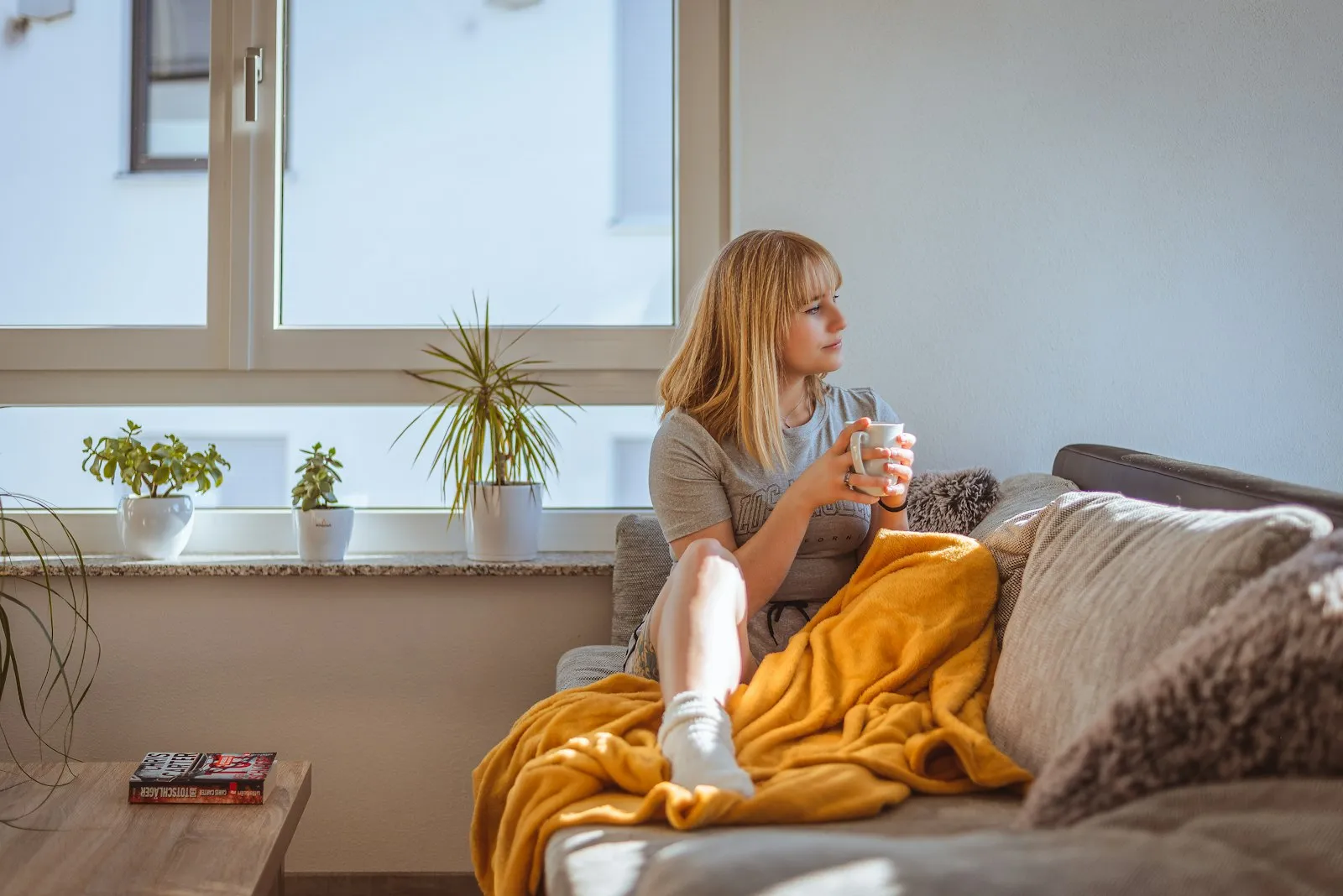 Une jeune femme se repose sur un canapé, une tasse à la main et un plaid jaune sur les jambes, près d'une fenêtre.